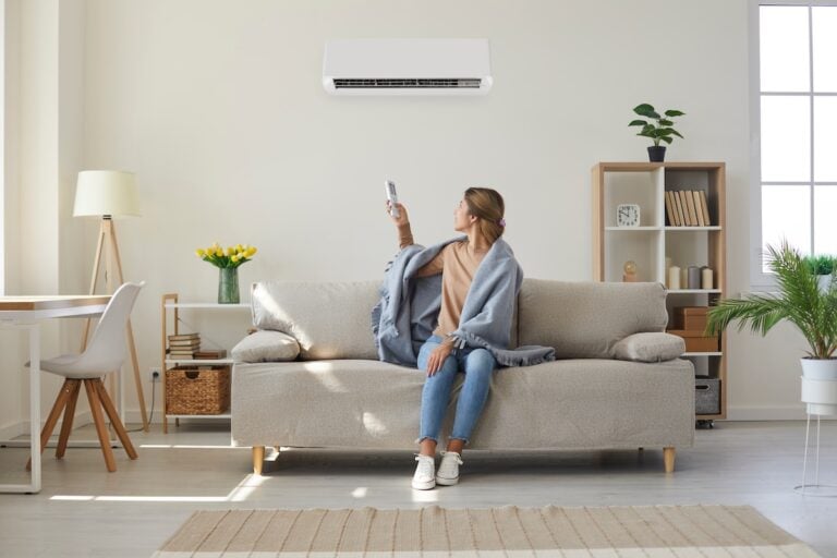 Woman who's sitting on sofa under warm plaid in living room switches off her air conditioner on wall. Young girl adjusting modern AC system, regulating temperature and enjoying cool fresh air at home
