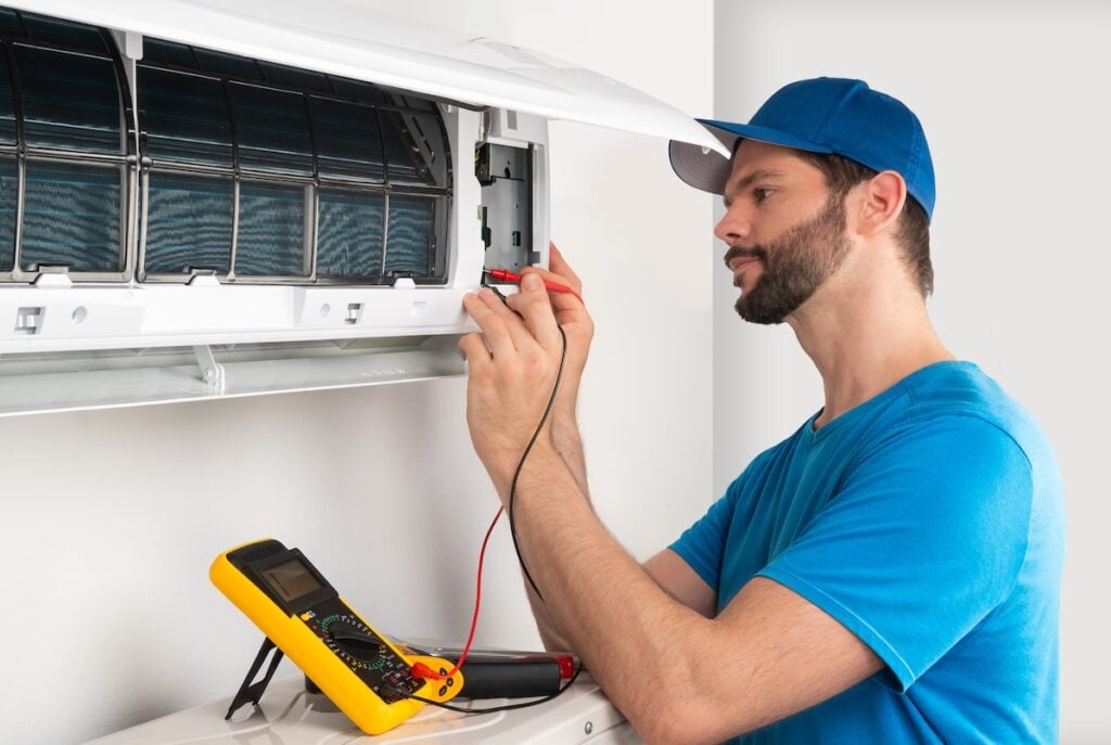 Installation service fix repair maintenance of an air conditioner indoor unit, by cryogenist technican worker with multimeter checking electric blue shirt and baseball cap