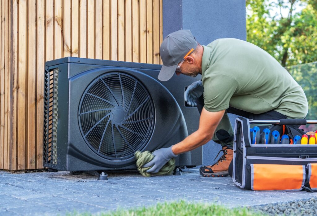 A technician in work attire cleans an outdoor air conditioning unit near a wooden wall. Tools are organized beside him for efficient service.