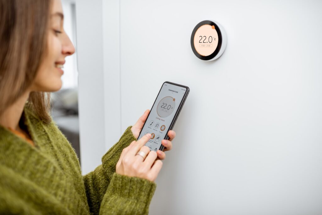 how to install smart thermostat Woman dressed in green sweater regulating heating temperature with a modern wireless thermostat and smart phone at home. Synchronization of thermostat with mobile devices concept